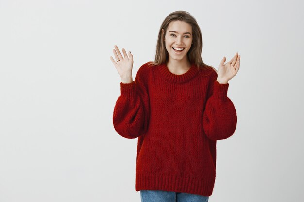 Girl telling friends about engagement. Portrait of excited pleased charming woman in beautiful red loose sweater, raising palms with ring on finger, cheering and expressing happiness over gray wall