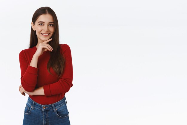 Girl thinking about interesing concept have good idea Attractive brunette woman in red blouse touching chin and smiling sly having creative plan standing thoughtful or pensive white background