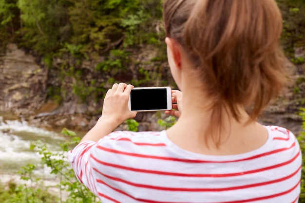 無料写真 少女旅行者は、スマートフォン、セレクティブフォーカス、エンジョイネイチャーズの風景、剥かれたシャツを着た女性、ポニーテールで渓谷の緑豊かな山川を撮ります。人と旅行のコンセプトです。