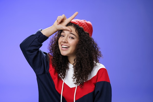 Girl triumphing and mocking friend as winning showing loser gesture on forehead and laughing having fun standing pleased and happy over blue background, smiling at camera joyfully.