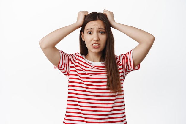 Girl in trouble holds hands on head, panicking. Alarmed young woman looking anxious and worried, distressed and frustrated with big problem, standing against white background