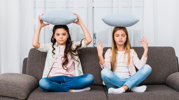 Ragazza che cerca di fare yoga come la sua amica mediando sul divano con cuscino