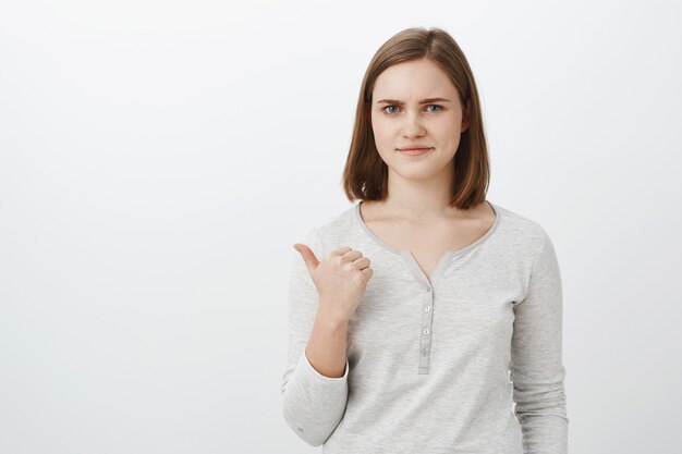 Girl unwilling bring lame classmate to party pointing backwards with thumb smirking displeased and dissatisfied rejection offer disapproving bad choice of friend standing unimpressed against grey wall