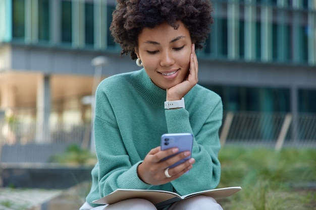 girl uses mobile phone takes break after learning material and preparing for exam poses with diary outdoors against urban setting wears casual jumper