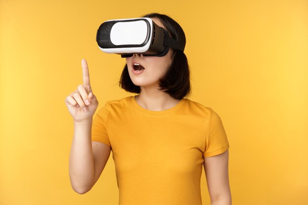 VRの女の子バーチャルリアリティメガネを使用して、黄色の背景の上に仮想的に立ってチャットをしている美しい若いアジアの女性