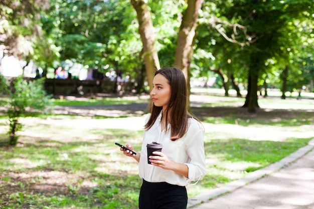 女の子は彼女の手の中で電話と公園でコーヒーの杯を歩く