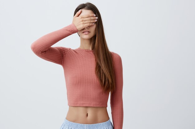 Girl don't want to see something. Close up of young beautiful caucasian female student in fashionable clothes clothing eyes with one hand. Body language.