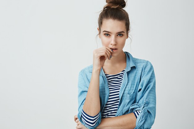 Girl wants to believe boyfriend but knows he is lying. Studio shot of suspicious focused woman with bun hairstyle, touching lips while thinking or hearing out excused, standing 