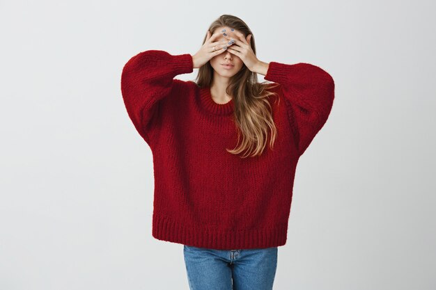 Girl wants to escape from world and reality. Studio portrait of stylish slender woman in red loose sweater covering eyes with both hands and standing calm and relaxed .