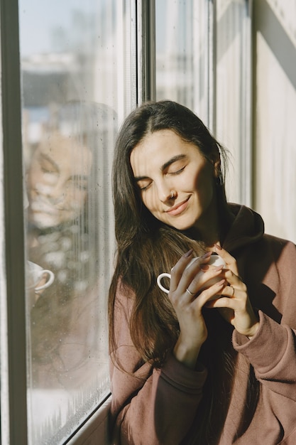 Foto gratuita ragazza in un cappotto caldo vicino alla finestra con una tazza di caffè