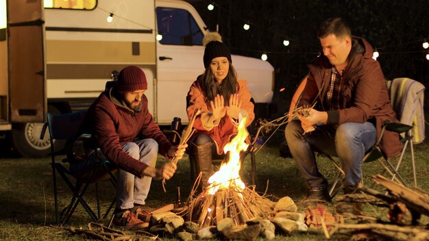 Girl warming up her hands while her friend making the fire stronger. Camp fire. Retro camper. Light bulbs.