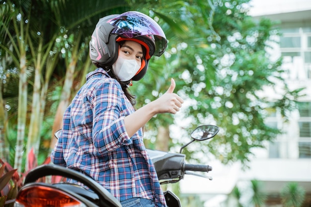 길가에서 오토바이에 엄지손가락으로 헬멧과 마스크를 쓴 소녀