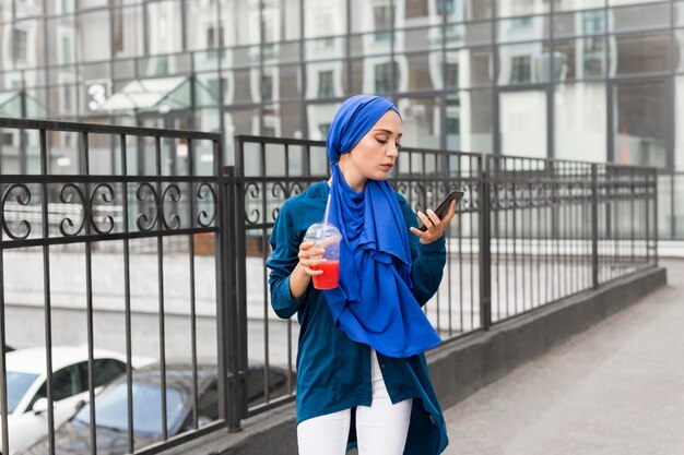 Девушка в хиджабе и держит смузи, глядя на свой телефон