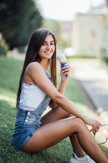 Girl wearing jeans combinaison sits on the grass outside in the city daytime summer icecream