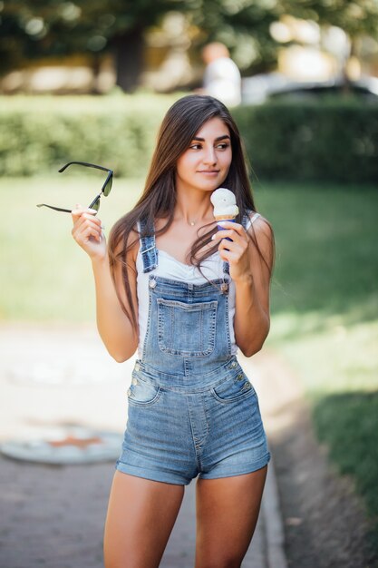 ジーンズの組み合わせとサングラスを身に着けている女の子は、街の昼間の夏に外でアイスクリームを食べる