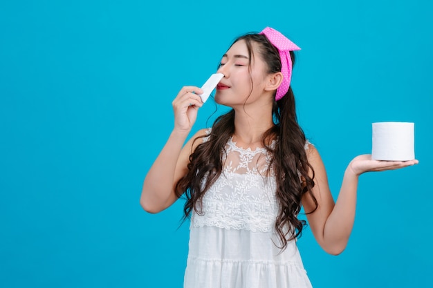 : A girl wearing a white pajamas sniffing a tissue and holding a tissue in her hand on a blue .