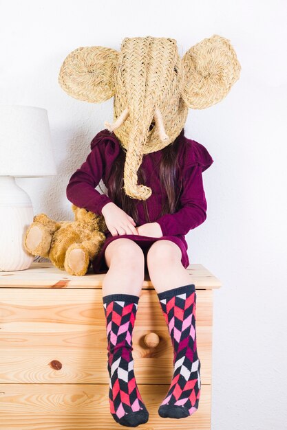 Девочка с плетеной маской слона, сидя на столе