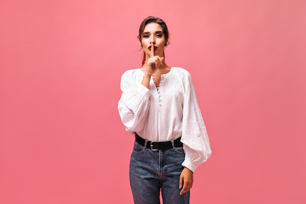 Girl in white blouse asking to keep secret. Gorgeous lady with big red lips in white long-sleeved shirt and jeans looks at camera.