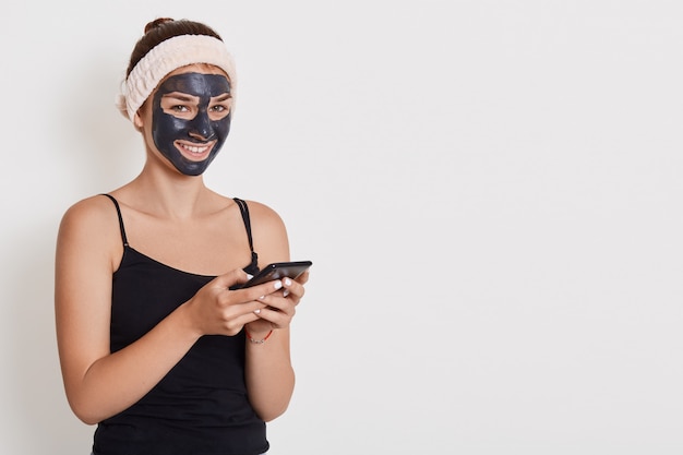 Girl in white hairband on her head and in clay black face mask holds mobile phone and writing message or reads news, doing beauty treatments at home, facial skin care.