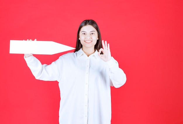 오른쪽을 가리키는 화살표를 들고 확인 손 기호를 보여주는 흰 셔츠에 소녀