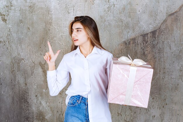 Girl in white shirt holding a pink gift box wrapped with white ribbon and having a good idea.