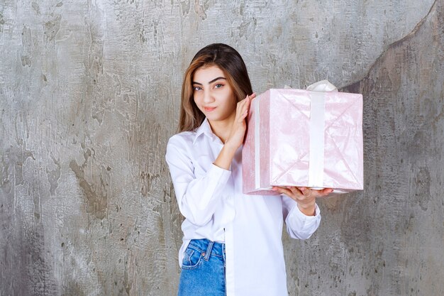 Girl in white shirt holding a pink gift box wrapped with white ribbon and looks confused and hesitating.
