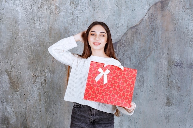 Girl in white shirt holding a red shopping bag and looks confused and thoughtful.