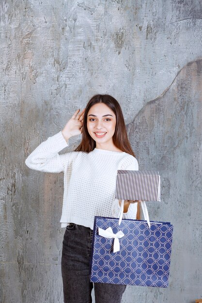 Girl in white shirt holding a silver gift box and a blue shopping bag and looking confused and thoughtful about making a choice.