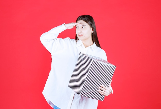 Free photo girl in white shirt holding a silver gift box, looking and calling someone