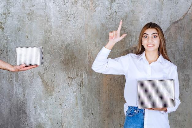 Girl in white shirt standing on a concrete wall is being offered a silver gift box and having a good idea.