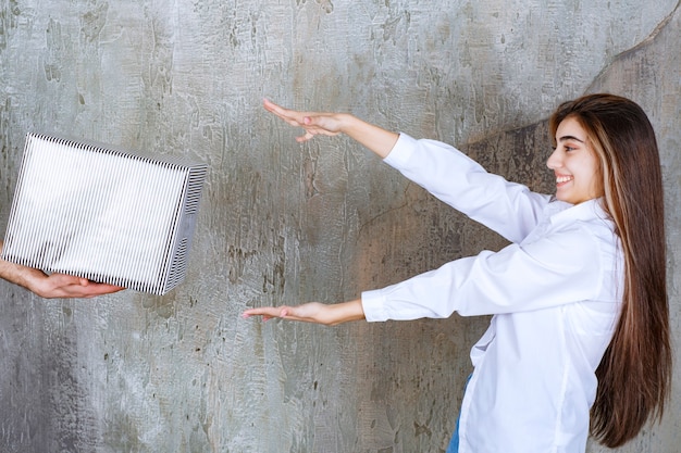 Foto gratuita alla ragazza in camicia bianca in piedi su un muro di cemento viene offerta una scatola regalo d'argento e le mani desiderose di prenderla