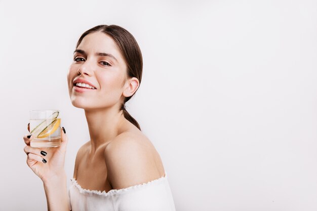 Girl in white top is smiling and posing on isolated wall. Portrait of brunette drinking healthy water with lemon and cucumber.