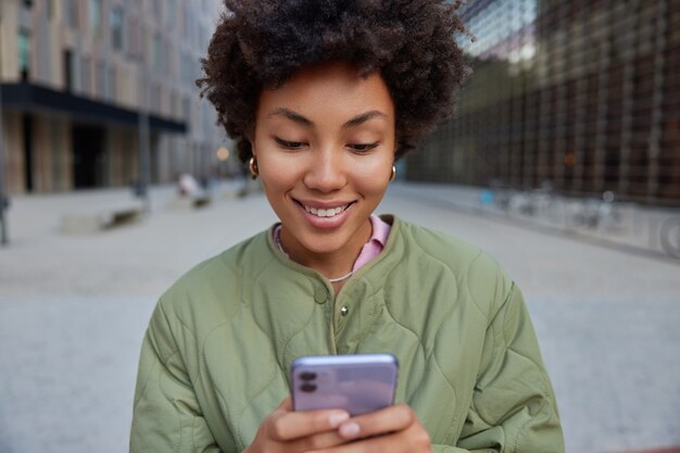 アフロの髪の少女がデジタルスマートフォンを持ってサーフィンソーシャルメディアがジャケットを着て笑顔が心地よく外でポーズをとってモビリティSMSを送信し、ネットワーキングサイトが街の通りに立っている