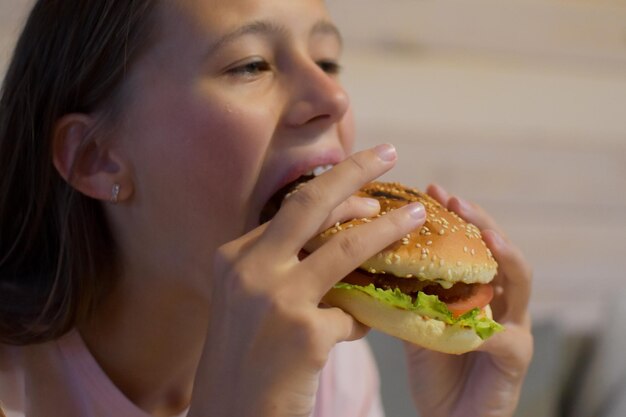 식욕이 있는 소녀는 맛있는 햄버거를 먹습니다. 아이가 큰 샌드위치 조각을 물다