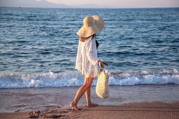 大きな帽子と籐のバッグを持った女の子が海岸を歩いています。夏休みのコンセプト。