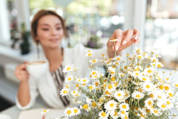 Girl with blond hair sitting in coffee shop with cup of cappuccino in hand and big bouquet of little chamomiles. Close up woman hand touching bouquet of chamomile flowers