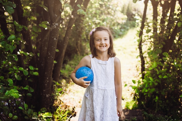Girl with blue ball stands in the rays of sun in forest 