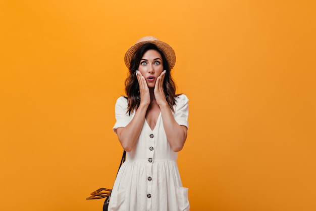 Girl with blue eyes stares in surprise at camera on orange background. Pretty woman with dark hair in straw hat and in white dress marvels.