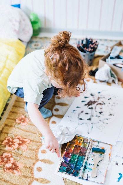 Girl with brush near set of water colors and paper sitting on floor