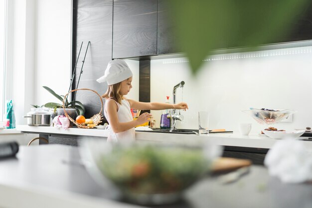 自宅の蛇口の下で彼女の手を洗うシェフの帽子を持つ少女