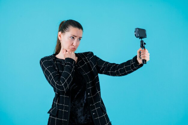 Girl with crying mimicry is taking selfie by putting hand under chin on blue background