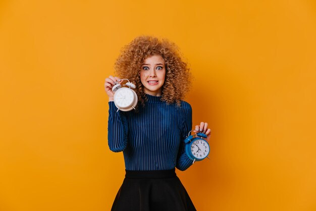 Girl with curls dressed in blue sweater feels awkward holding two alarm clocks on yellow background