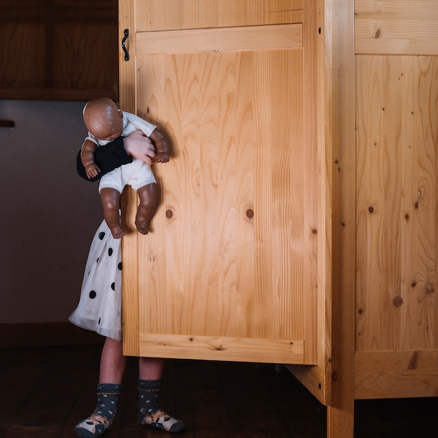 Girl with doll standing behind wooden cupboard