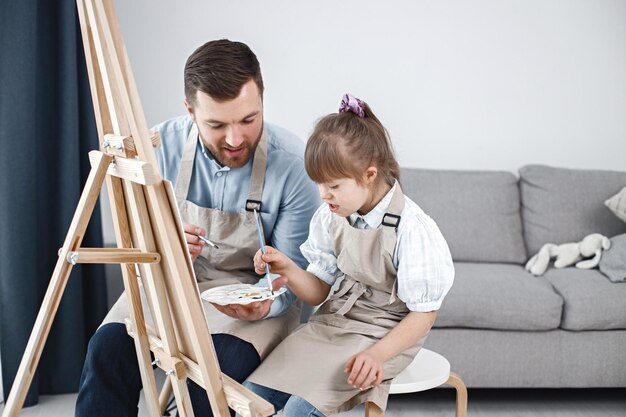 Девочка с синдромом Дауна и ее отец рисуют на мольберте кистями