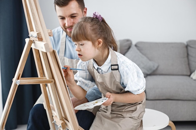 다운 증후군을 앓고 있는 소녀와 그녀의 아버지는 붓으로 이젤에 그림을 그립니다.