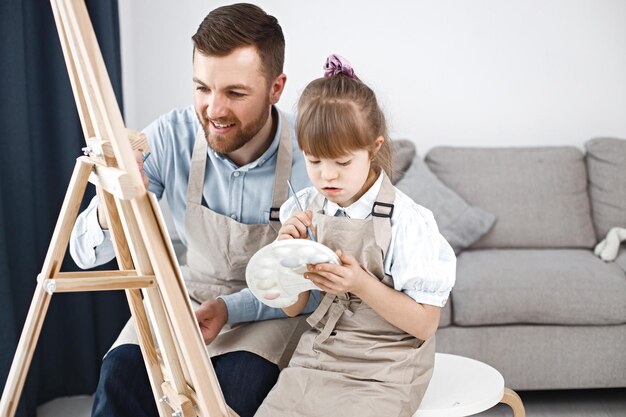 다운 증후군을 가진 소녀와 그녀의 아버지는 붓으로 이젤에 그림을 그립니다.