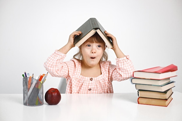 Free photo girl with down syndrome sitting at desk and holding a book on her head