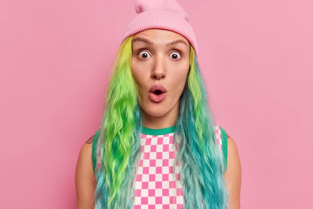 Free photo girl with dyed long hair piercing in nose gazes surprisingly at camera keeps mouth opened wears hat checkered dress isolated on pink