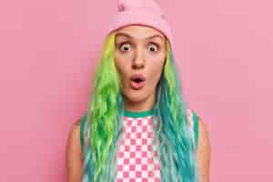 Free photo girl with dyed long hair piercing in nose gazes surprisingly at camera keeps mouth opened wears hat checkered dress isolated on pink