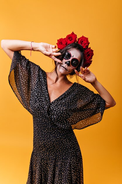 Girl with ethnic Mexican face mask posing with mysterious look on isolated wall. Portrait of unusual woman with roses in her hair 